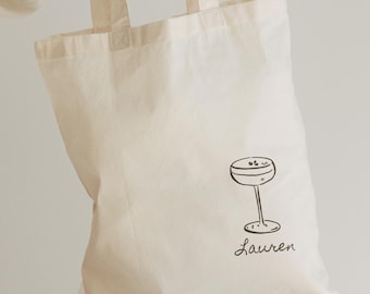 May include: Cream-colored tote bag with a black illustration of a cocktail glass and the name "Lauren" in cursive. The bag has two handles and is held up by a person.