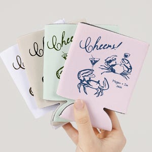 May include: A selection of drink koozies in white, beige, mint green, and pink. The pink koozie features the word "Cheers!" and a crab illustration with the names "Megan + Joe 2024". The other koozies have the word "Cheers" in a script font.