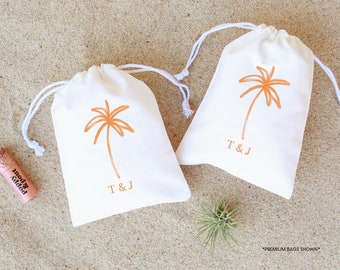 May include: Two white cotton drawstring bags with orange palm tree designs and the initials "T & J" printed on the front. The bags are sitting on a sandy surface.