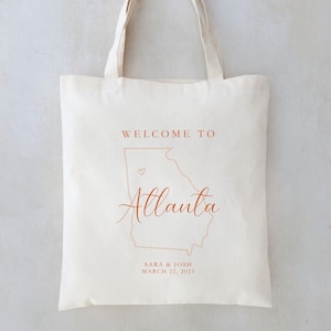 May include: White canvas tote bag with brown text that says "Welcome to Atlanta" with an outline of the state of Georgia and a small heart. The text "Sara & Josh March 22, 2025" is also printed on the bag.