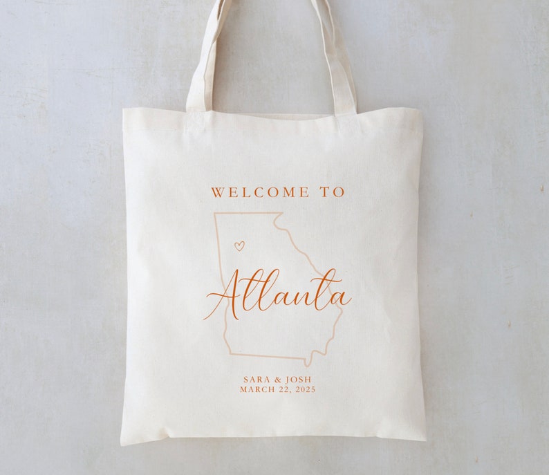 May include: White canvas tote bag with brown text that says "Welcome to Atlanta" with an outline of the state of Georgia and a small heart. The text "Sara & Josh March 22, 2025" is also printed on the bag.