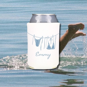 May include: A white can cooler with a blue line drawing of swimsuits hanging on a clothesline. The name "Emmy" is written in cursive below the drawing. The background is a blurred image of water.