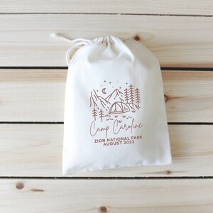 May include: A white drawstring bag with a brown illustration of a campsite with mountains, trees, a tent, and stars. The text "Camp Caroline" is written below the illustration, followed by "Zion National Park August 2023".