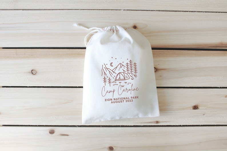 May include: A white drawstring bag with a brown illustration of a campsite with mountains, trees, a tent, and stars. The text "Camp Caroline" is written below the illustration, followed by "Zion National Park August 2023".