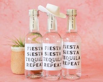 May include: Three clear glass bottles with white labels that say "Fiesta Siesta Tequila Repeat" on a pink background. A white cowboy hat sits on top of the middle bottle.