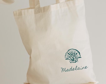 May include: Cream-colored tote bag with a teal seashell graphic and the name "Madelaine" in a script font. The bag has two handles and is made of a soft, natural fabric. The bag is sitting on a white surface.