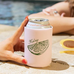 May include: A light pink can cooler with the name "Kelsey" and a green grapefruit slice design. The can cooler holds a silver can of grapefruit soda. The background shows a pool and a person.