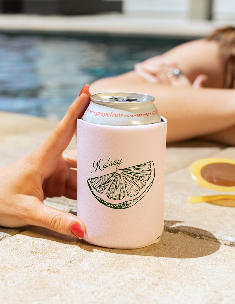 May include: A light pink can cooler with the name "Kelsey" and a green grapefruit slice design. The can cooler holds a silver can of grapefruit soda. The background shows a pool and a person.