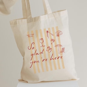 May include: Cream-colored tote bag with a design featuring vertical stripes and red illustrations of a wine glass, sandals, and a seashell. The text "So glad you're here" is written in red script. The bag also includes the text "Jadyn + Noah Italy 2026".