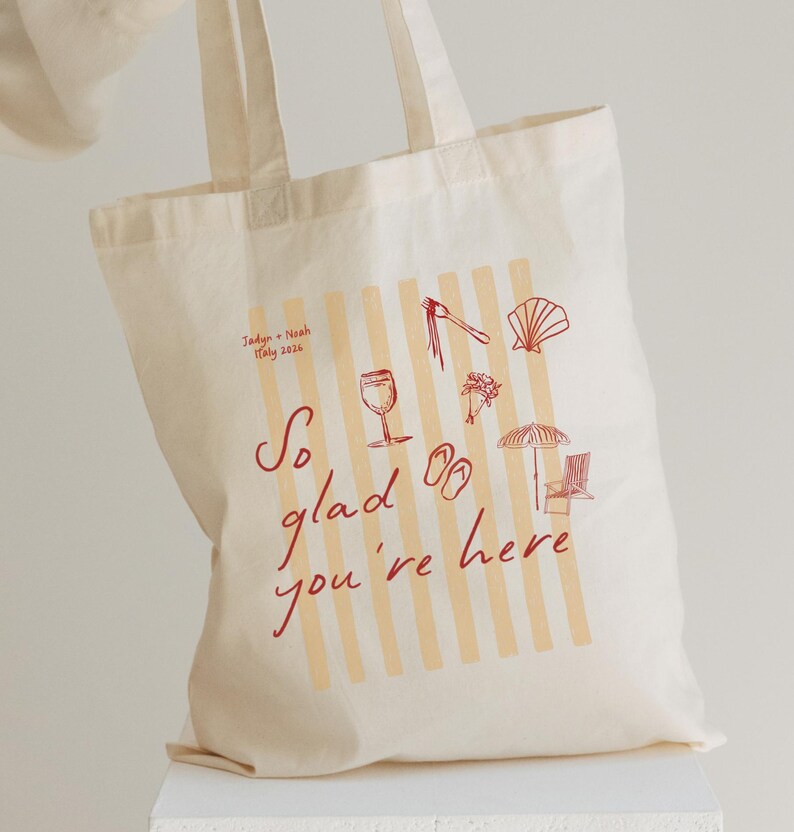 May include: Cream-colored tote bag with a design featuring vertical stripes and red illustrations of a wine glass, sandals, and a seashell. The text "So glad you're here" is written in red script. The bag also includes the text "Jadyn + Noah Italy 2026".