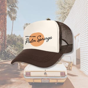 Pu&ograve; includere: Un cappello da camionista bianco e marrone con una rete marrone sul retro. La parte anteriore del cappello ha un cerchio arancione con il testo "With love from Palm Springs" in nero.