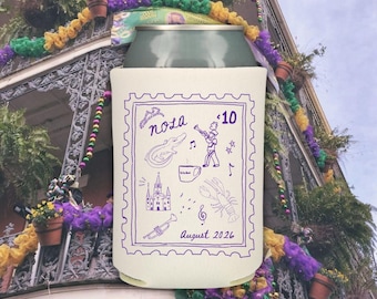 May include: A cream-colored can cooler with a purple stamp-like design featuring the word "nola", an alligator, a musician, and other New Orleans icons. The text "August 2026" is also included.