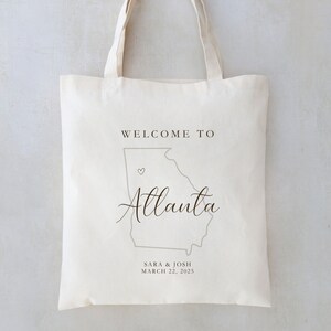 May include: White canvas tote bag with a gray outline of the state of Georgia and the text "Welcome to Atlanta" in black cursive font. A small heart is placed on the outline of Atlanta. The text "Sara & Josh March 22, 2025" is printed below the outline of Georgia.