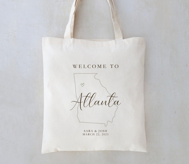 May include: White canvas tote bag with a gray outline of the state of Georgia and the text "Welcome to Atlanta" in black cursive font. A small heart is placed on the outline of Atlanta. The text "Sara & Josh March 22, 2025" is printed below the outline of Georgia.