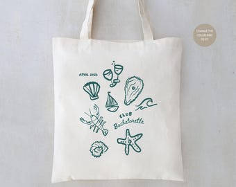 May include: A white canvas tote bag with a teal design featuring a variety of nautical and beach-themed illustrations, including a sailboat, seashells, an oyster, a lobster, a starfish, and two wine glasses. The text "APRIL 2023" and "CLUB Bachelorette" are also included in the design.
