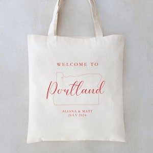 May include: A white tote bag with red text that says "Welcome to Portland" with an outline of the state of Oregon and a small heart. The text "Aliana & Matt July 2024" is also printed on the bag.