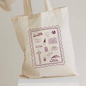 Peut inclure: Sac fourre-tout crème avec un motif violet en forme de timbre représentant les monuments de Seattle et le texte "Seattle" et "Laura's Bachelorette". Le motif comprend des illustrations d'un avion, d'un poisson, de la Space Needle et d'une baleine.