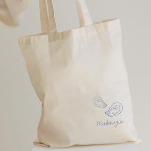 May include: Cream-colored tote bag with two blue oyster illustrations and the name "Makenzie" printed in a matching blue font. The bag has two handles and is made of a sturdy fabric.