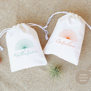 May include: Two white cotton drawstring bags with a sunburst design and personalized names. The bag on the left has a teal sunburst and the name "Katherine". The bag on the right has an orange sunburst and the name "Caroline".