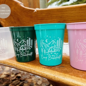 May include: Four plastic cups with a white mountain and tent design and the text "Camp Bachelorette". The cups are in the colors clear, green, teal, and pink.