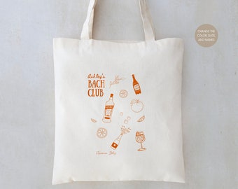 May include: A white canvas tote bag with an orange design featuring the text "Ashley's Bach Club" and illustrations of drinks, fruit, and a sprig of rosemary. The text "Florence, Italy" is printed at the bottom of the design.