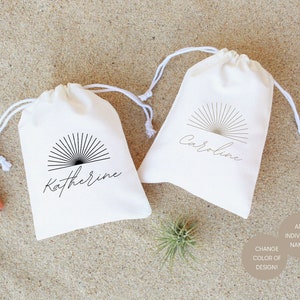 May include: Two white cotton drawstring bags with a black sunburst design and personalized names, "Katherine" and "Caroline". The bags are on a sandy surface with a small air plant and a tube of lip balm.