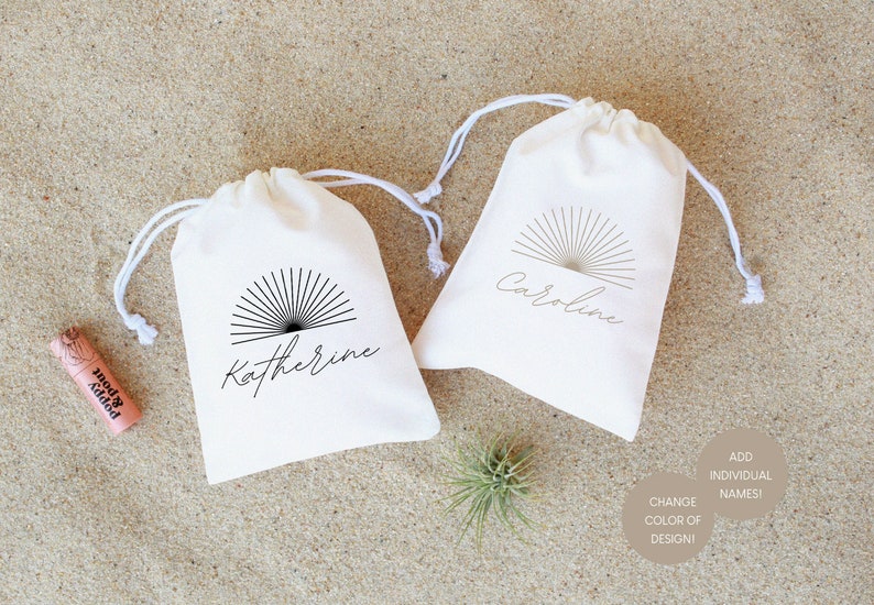 May include: Two white cotton drawstring bags with a black sunburst design and personalized names, "Katherine" and "Caroline". The bags are on a sandy surface with a small air plant and a tube of lip balm.