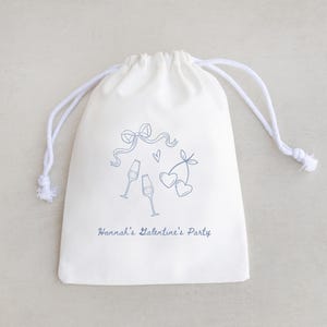 Pu&ograve; includere: Una borsa di cotone bianco con coulisse, con un design blu di bicchieri di champagne, cuori e un fiocco. Il testo "Hannah's Valentine's Party" &egrave; stampato sulla borsa.