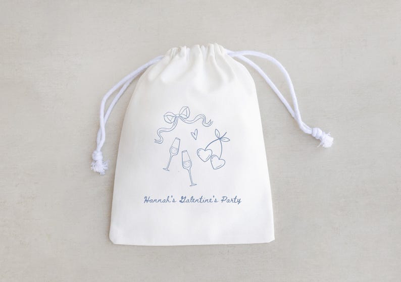 Pu&ograve; includere: Una borsa di cotone bianco con coulisse, con un design blu di bicchieri di champagne, cuori e un fiocco. Il testo "Hannah's Valentine's Party" &egrave; stampato sulla borsa.