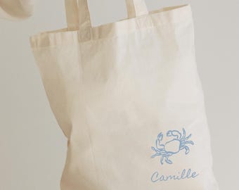 May include: Cream-colored canvas tote bag with a light blue crab illustration and the name "Camille" printed below. The bag has two handles and is made of a sturdy fabric, perfect for carrying groceries or personal items.