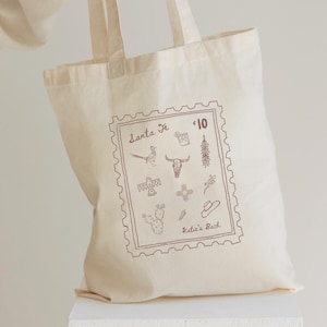 May include: Cream-colored tote bag with a brown stamp-like design featuring the text "Santa Fe" and various Southwestern-themed illustrations. The bag has two handles and the text "Katie's Bach".