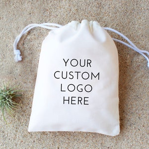 May include: White cotton drawstring bag with the text "YOUR CUSTOM LOGO HERE" printed in black.