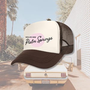 Pu&ograve; includere: Un cappello da camionista bianco e marrone con un cerchio rosa e il testo "With love from Palm Springs" in nero.