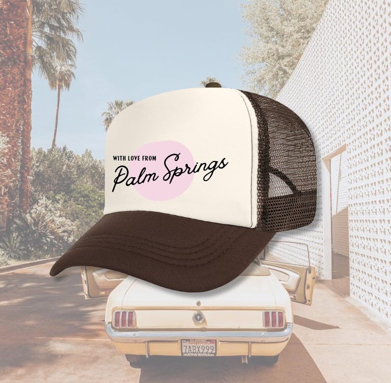 Pu&ograve; includere: Un cappello da camionista bianco e marrone con un cerchio rosa e il testo "With love from Palm Springs" in nero.