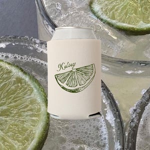 May include: A cream-colored can cooler with the name "Kelsey" and a lime slice graphic in green. The cooler is on a can, with a background of drinks with lime slices and ice.