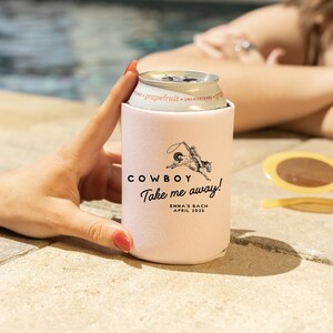 May include: Pink can cooler with a black and white illustration of a cowboy riding a horse. The text "COWBOY Take me away!" is printed on the cooler. Below the text is "EMMA'S BACH APRIL 2023".