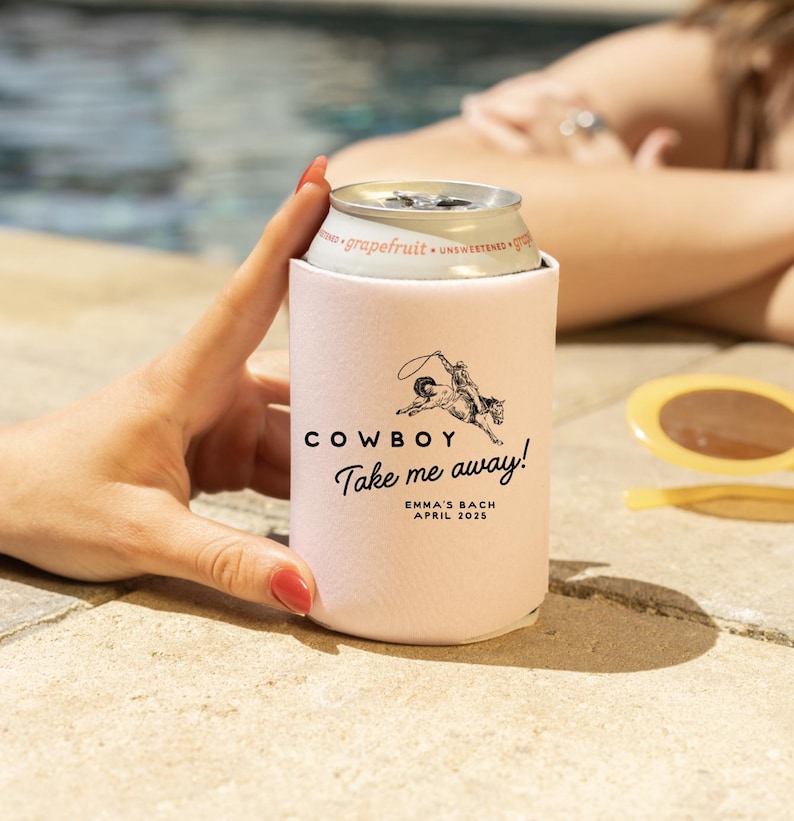 May include: Pink can cooler with a black and white illustration of a cowboy riding a horse. The text "COWBOY Take me away!" is printed on the cooler. Below the text is "EMMA'S BACH APRIL 2023".