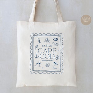 May include: A white tote bag with a blue stamp design featuring the text "CAPE COD" and the date "09.21.24". The stamp also includes various nautical icons like a lighthouse, sailboat, anchor, seashells, and a ring. The text "Madeline and Eli" is also included.