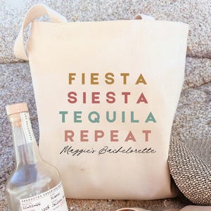 May include: A white canvas tote bag with the text "FIESTA SIESTA TEQUILA REPEAT" in colourful block letters. The text "Maggie's Bachelorette" is written in cursive below the block letters.
