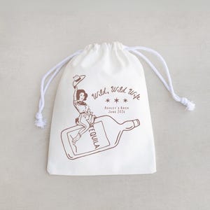 May include: A white drawstring bag with a brown illustration of a woman riding a tequila bottle. The text "Wild, Wild Wife" and "Ashley's Bach June 2016" are also printed on the bag. The bag has white drawstrings.