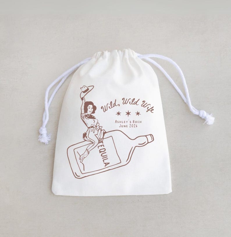 May include: A white drawstring bag with a brown illustration of a woman riding a tequila bottle. The text "Wild, Wild Wife" and "Ashley's Bach June 2016" are also printed on the bag. The bag has white drawstrings.