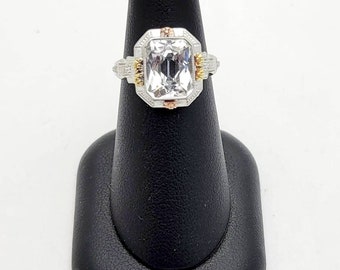 May include: A silver and rose gold ring with a large, clear, rectangular-shaped gemstone. The gemstone is surrounded by smaller diamonds and the band is set with smaller diamonds.