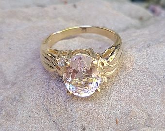May include: A gold-toned ring with an oval, light pink gemstone. The ring has a detailed band with a swirling design and small accent stones on each side of the main stone. The ring is set on a textured, light-colored surface.