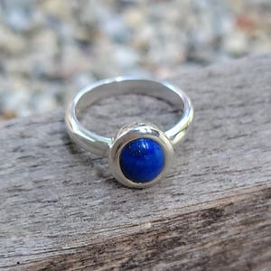 May include: A silver ring featuring a smooth, round, deep blue lapis lazuli gemstone set in a polished metal bezel. The band is plain and metallic, showcasing a simple design that highlights the vibrant color of the circular stone at the center.