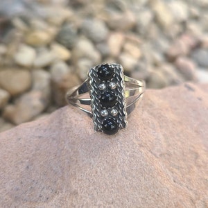 May include: A silver ring with a rectangular setting featuring four black gemstones. The ring has a delicate, intricate design with small beads around the edges of the setting.