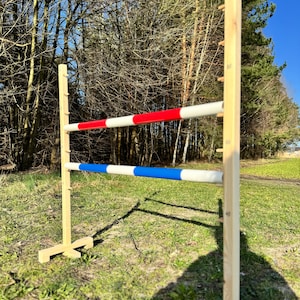 May include: A wooden show jumping obstacle with two coloured poles. The top pole is red and white, the bottom is blue and white. The uprights have height markers in centimetres and are set on grass.