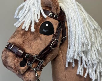 May include: A plush toy horse head in shades of brown with a white mane and tail. It features a brown leather bridle with silver-colored hardware and black eye details. The toy is designed to be held like a stick horse.
