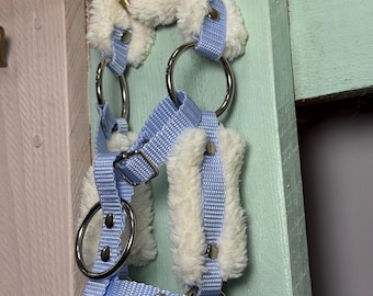 May include: A light blue and white restraint with fluffy white padding and silver metal rings. The restraint is made of light blue nylon webbing and is designed to be adjustable. The fluffy padding is soft and comfortable.