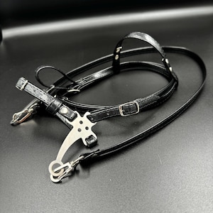 May include: Black leather camera strap with a metal buckle and a silver metal hook. The strap is designed to be worn around the neck or shoulder.