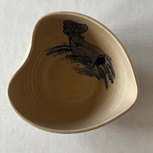 May include: A beige ceramic bowl with an asymmetrical shape. The interior features a hand-painted illustration of a figure in a long dress and hat, rendered in black and gray. The bowl has a smooth, glossy finish.
